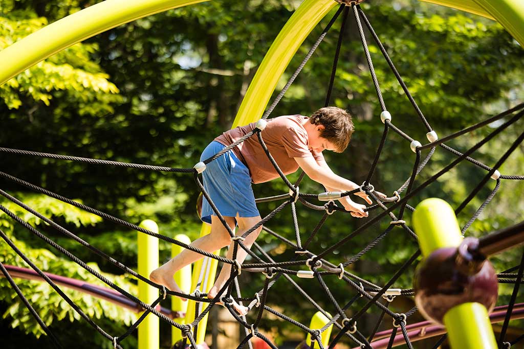Young boy climbs on playground structure nestled in the woods.